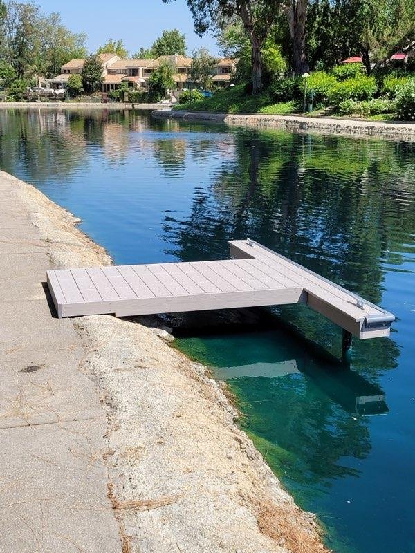 A lakeside dock extending over the water with a boat lift at the end, surrounded by trees, houses, and a clear blue sky.