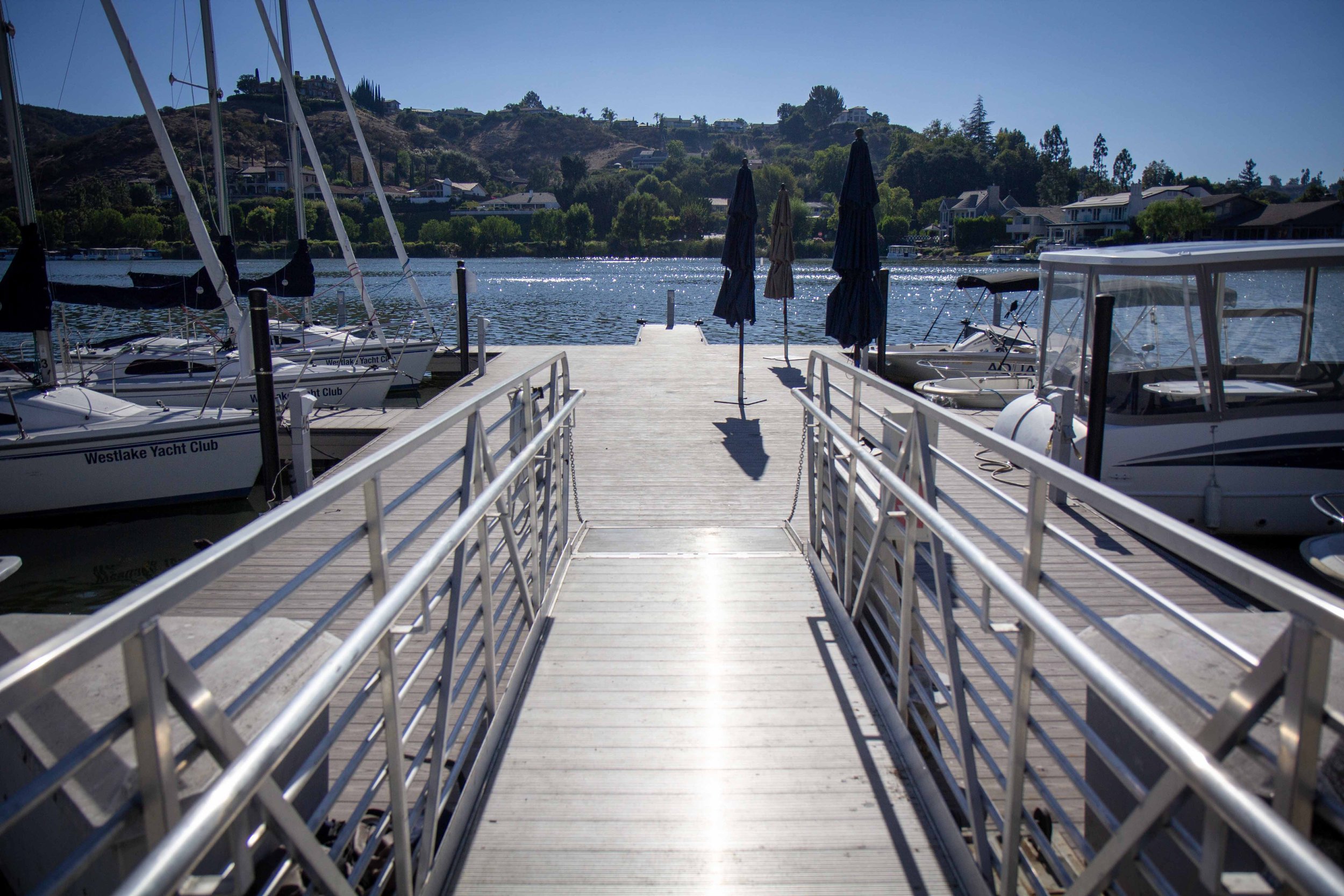 A dock leading to the water with boats on either side, some with sails, and a cityscape with houses and trees across the water.