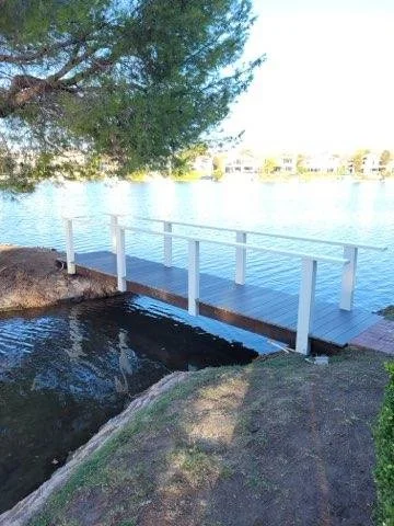 A small wooden dock extending over a body of water, with a white railing along the edge, near rocks and a tree.