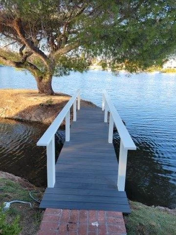 A small dock with white railings extending over a lake, with a large tree on the left and a brick path in front.