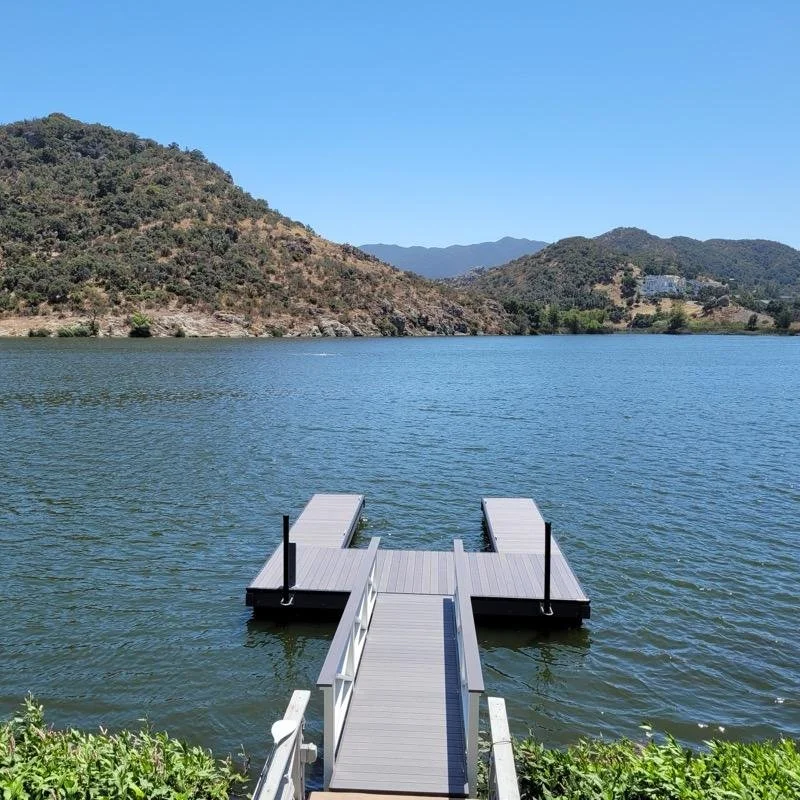 A dock extends into a calm lake, surrounded by hills and mountains under a clear blue sky.