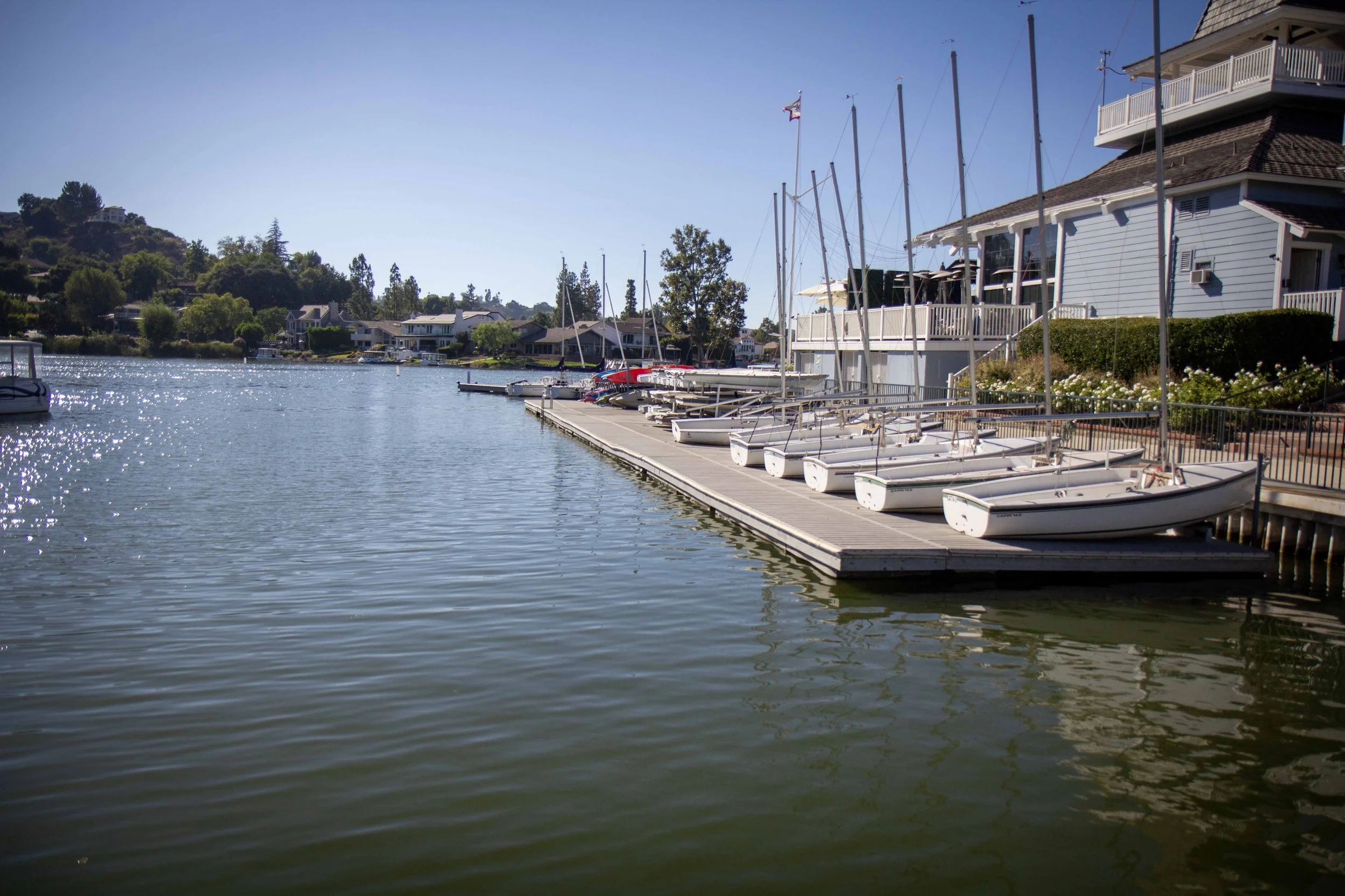 Sailing boats docked along a waterfront marina with houses and trees in the background under a clear blue sky.