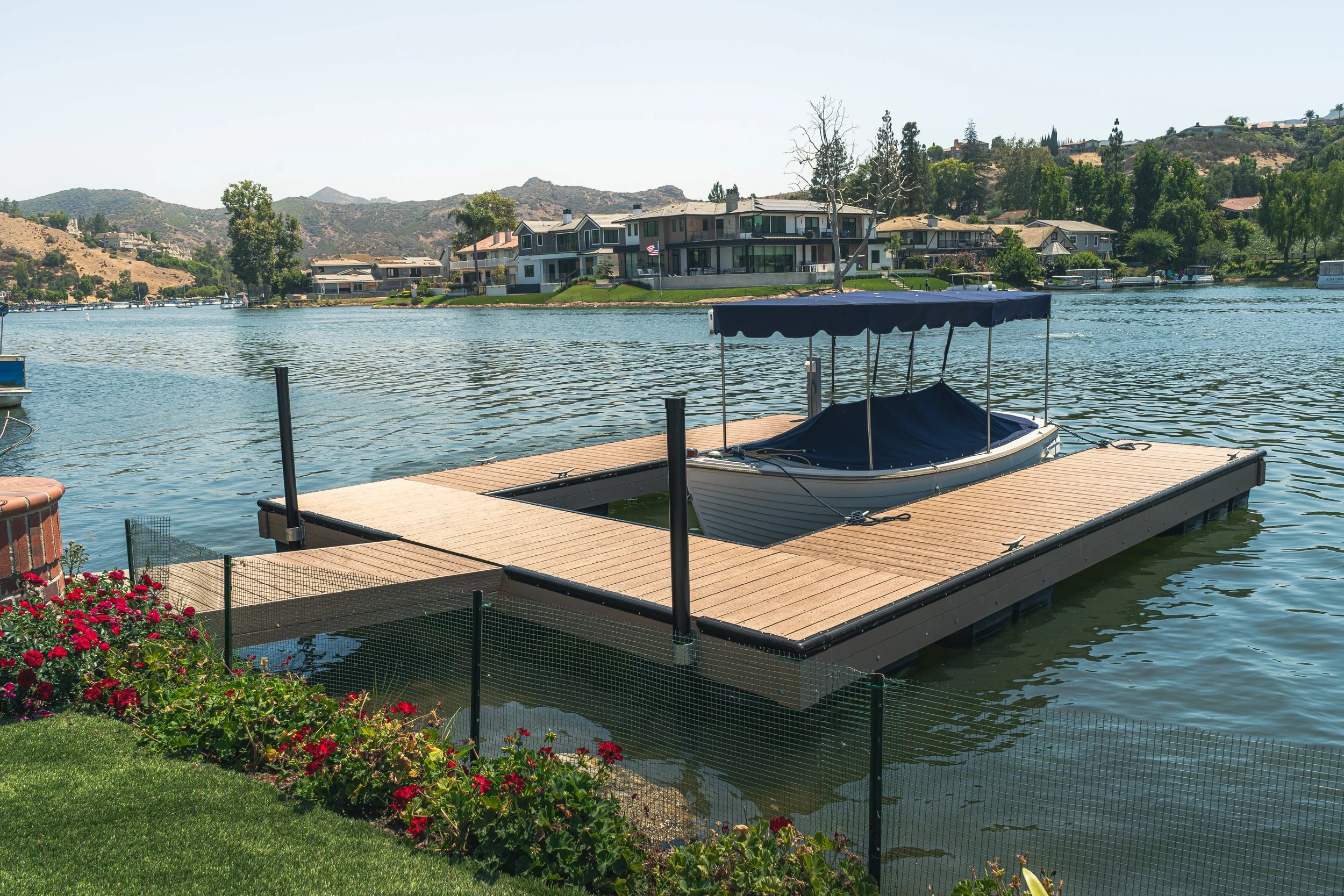 A wooden dock with a boat covered with a dark blue tarp, floating on a river with residential houses and hills in the background.
