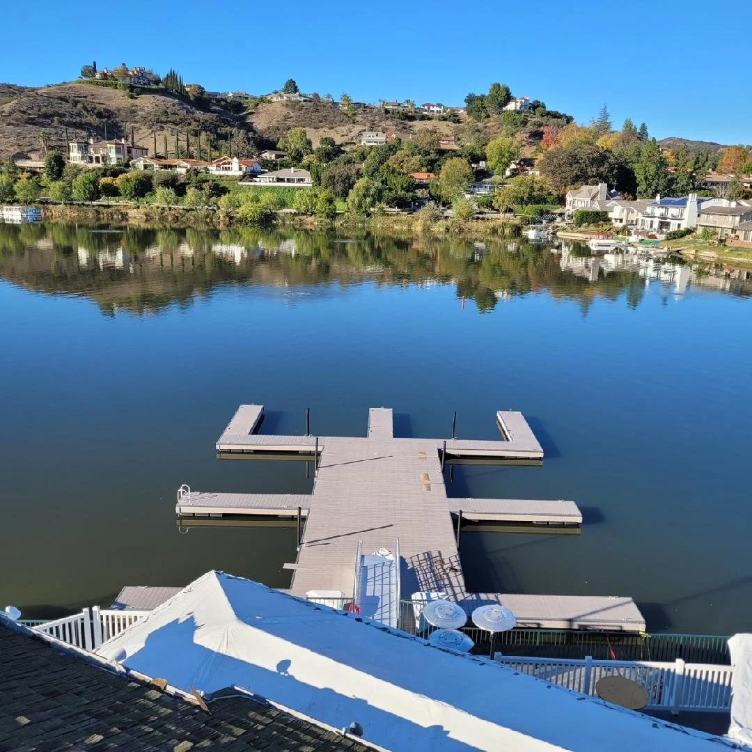 A canalside dock extending into a calm body of water with houses and wooded hill in the background.