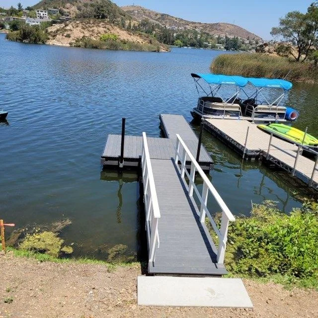 A marina with a dock extending into a lake, featuring boats including a pontoon boat with a blue canopy, and a kayak on a dock, surrounded by hilly terrain and trees.