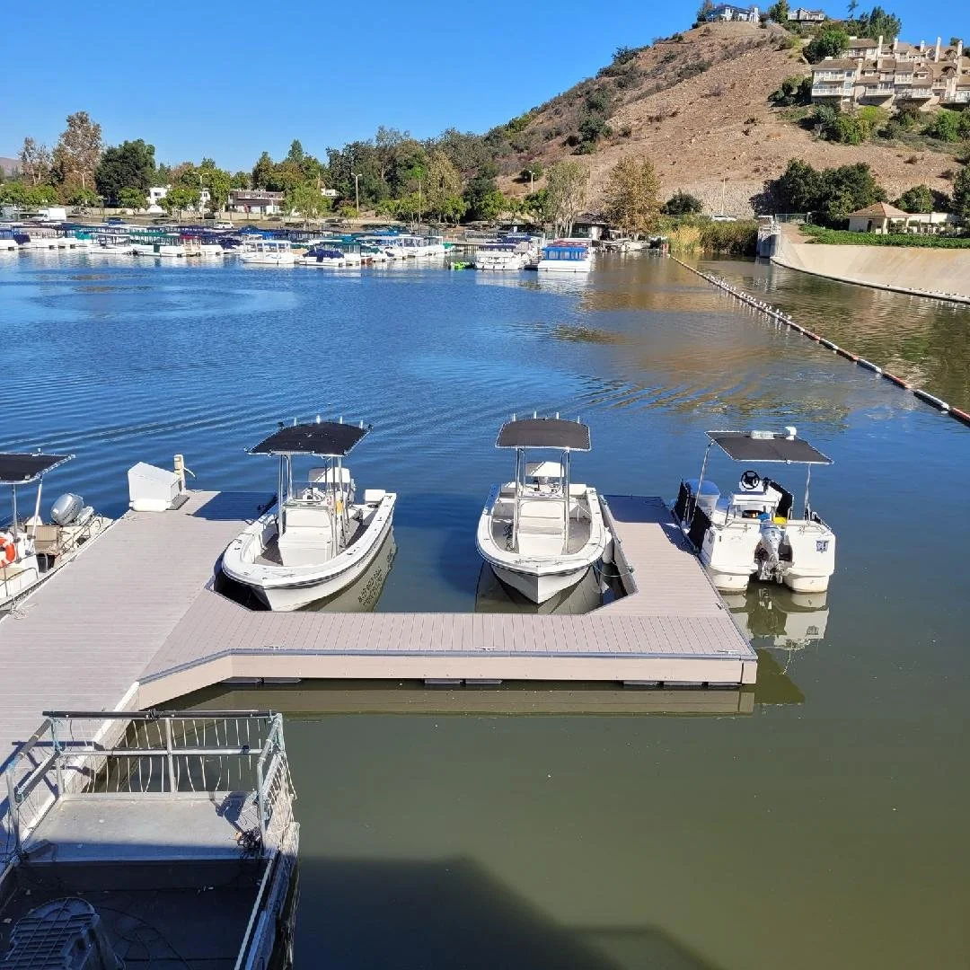 Marina with three boats docked at a floating pier on a calm lake, with a hillside in the background and other boats lined up along the shore.