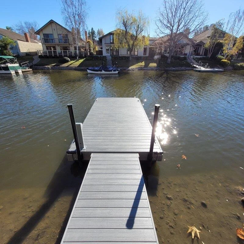 A wooden dock extends into a river with houses and boats along the opposite shoreline. Bare trees line the riverbank, and the sun reflects off the water.