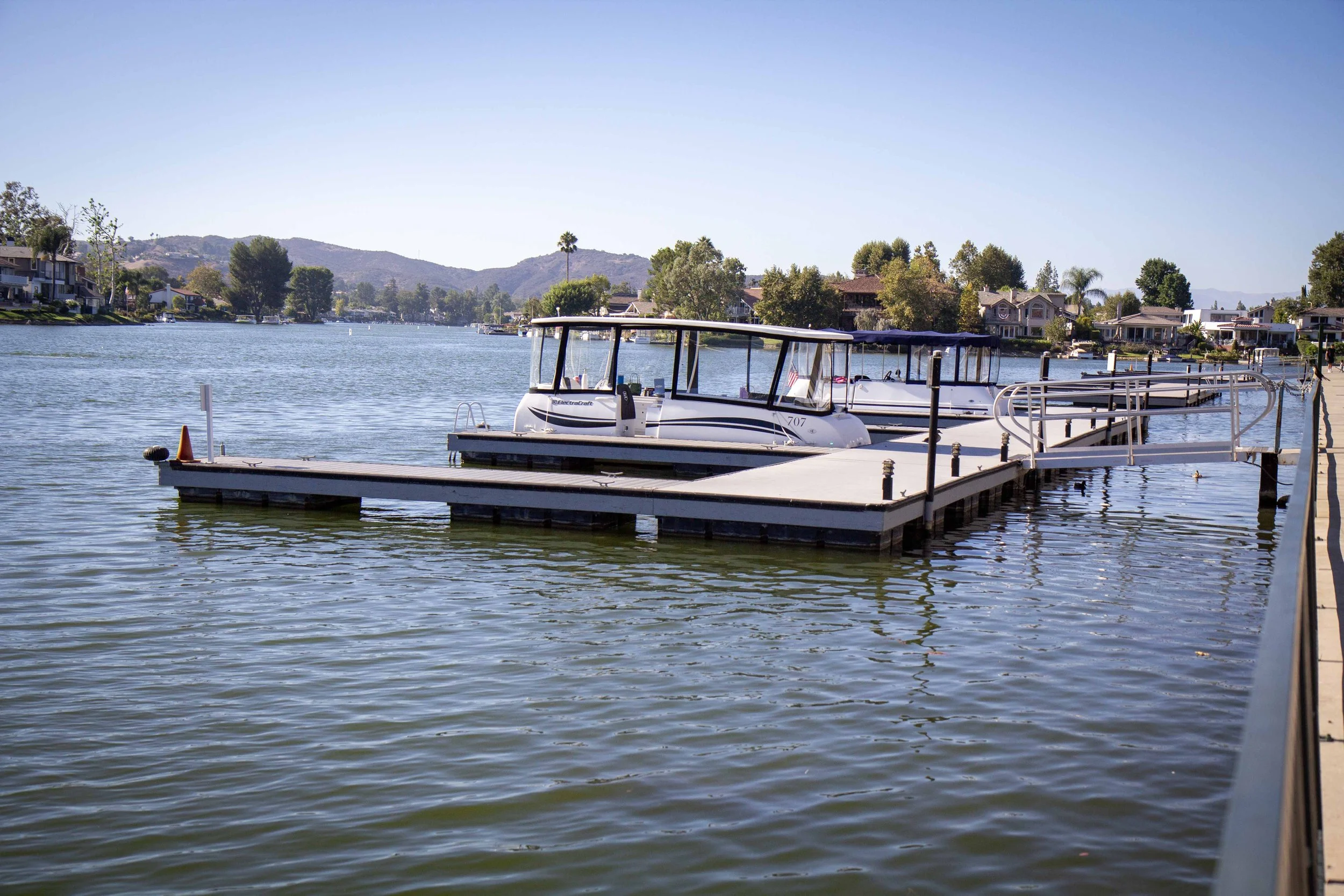Boats docked at a marina on a calm lake with houses and hills in the background during a sunny day.