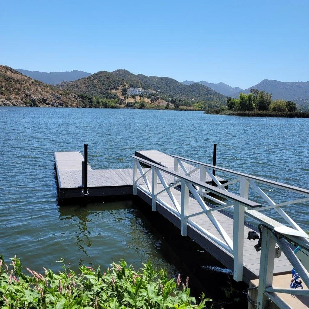A wooden dock extends into a lake with mountains in the background and clear blue sky above.