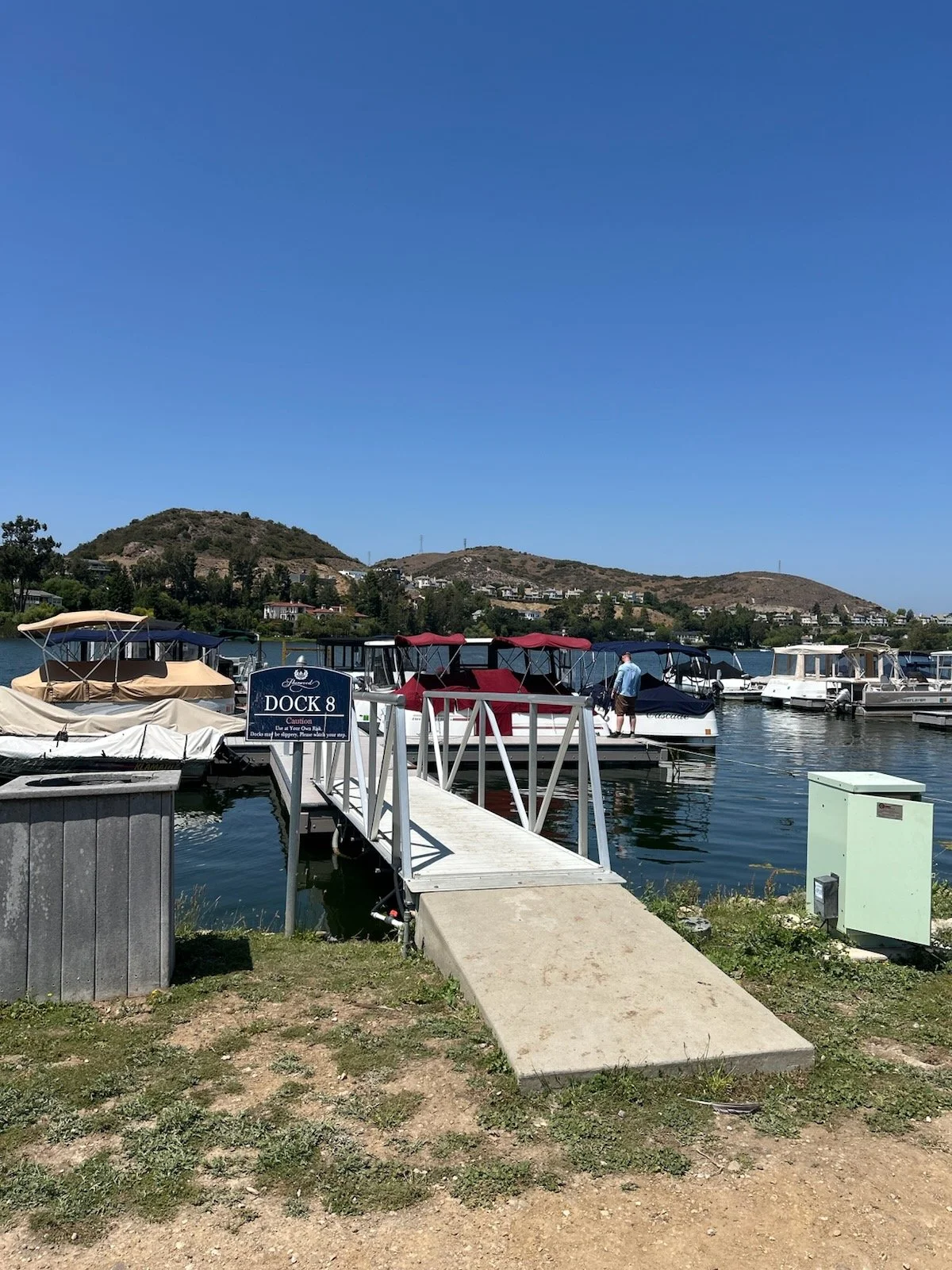 Dock 8 at a marina with several boats moored on calm water, a pedestrian ramp leading onto the dock, a blue sign, and hilly landscape in the background under a clear blue sky.