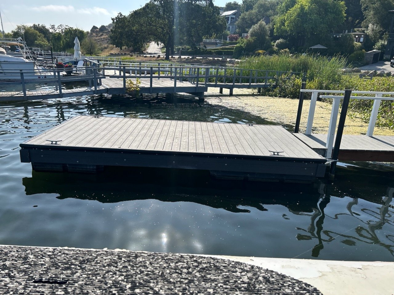 Dock floating on a body of water, with other docks and boats in the background, surrounded by trees and residential buildings.