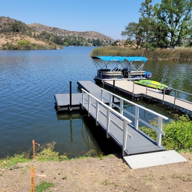 A dock extending into a creek with a boat, kayak, and paddleboard on it, surrounded by trees and hills under a clear blue sky.