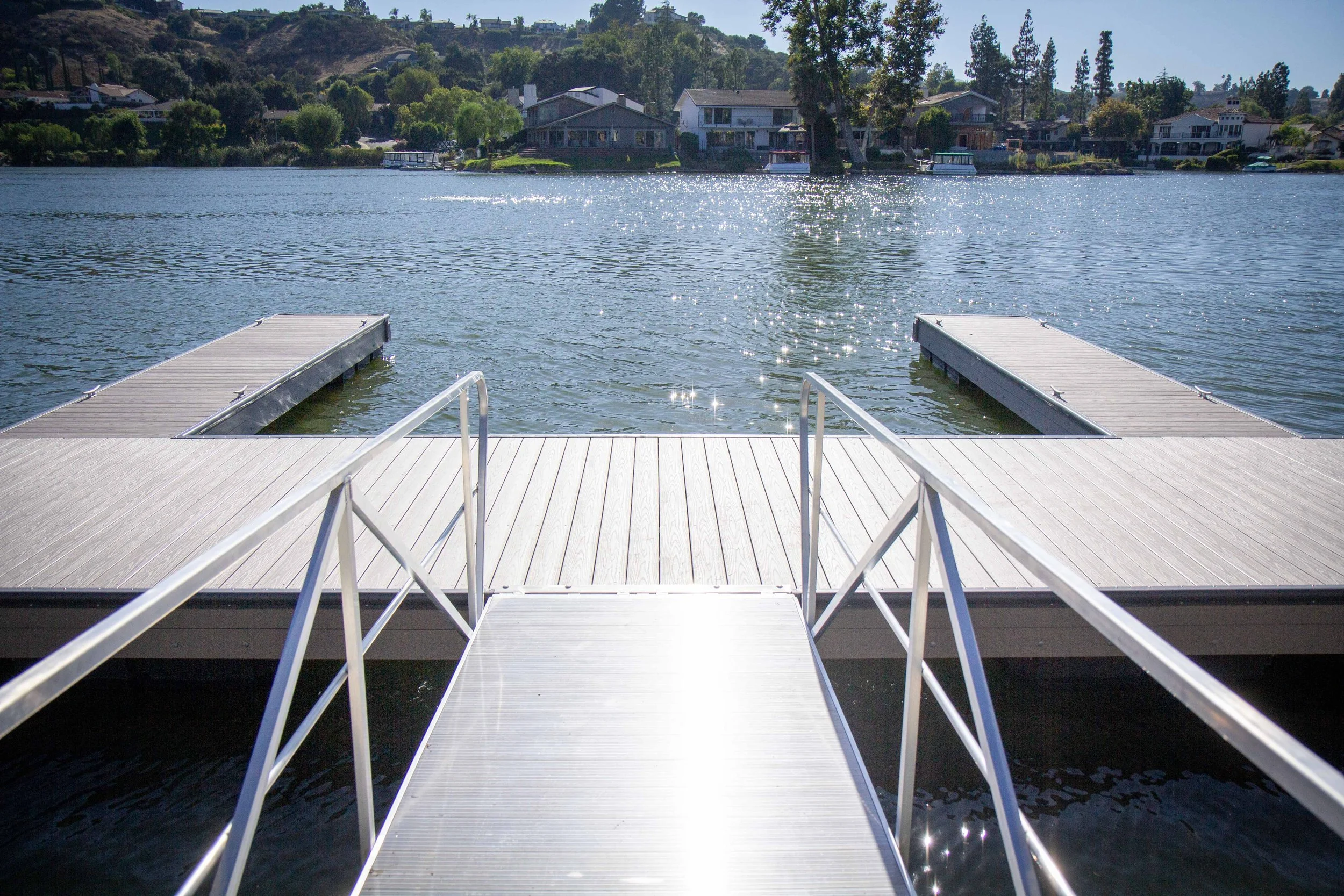 A lakeside dock with two floating platforms extends into the water, with houses and trees on the opposite shore under a clear sky.