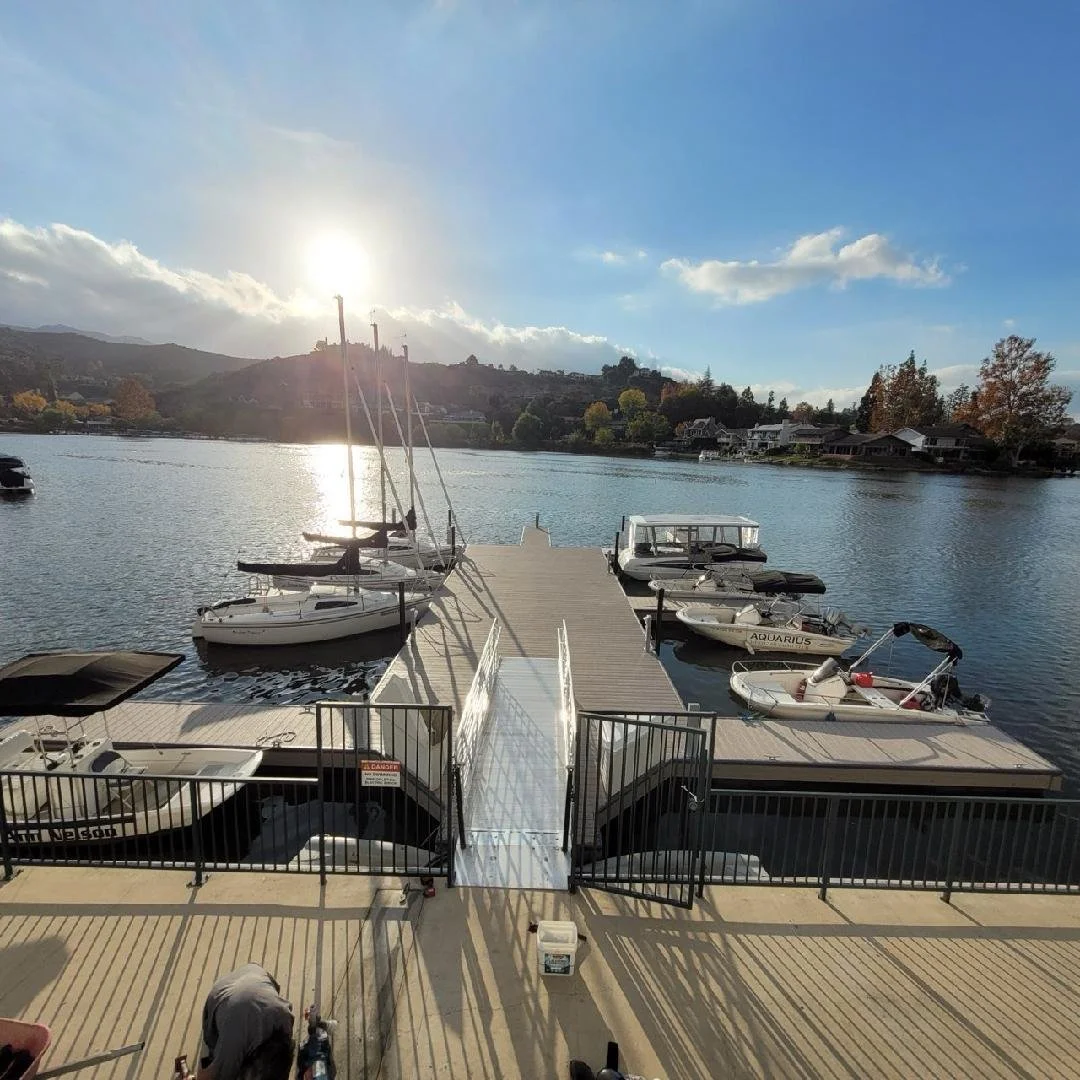 A dock with boats moored, overlooking a calm body of water and a hillside with houses and trees in the background, under a partly cloudy sky with the sun shining.