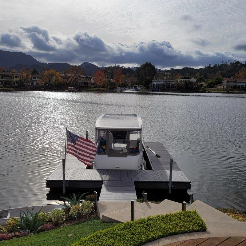 A dock with a boat and an American flag on a lakeside property with houses and mountains in the background.