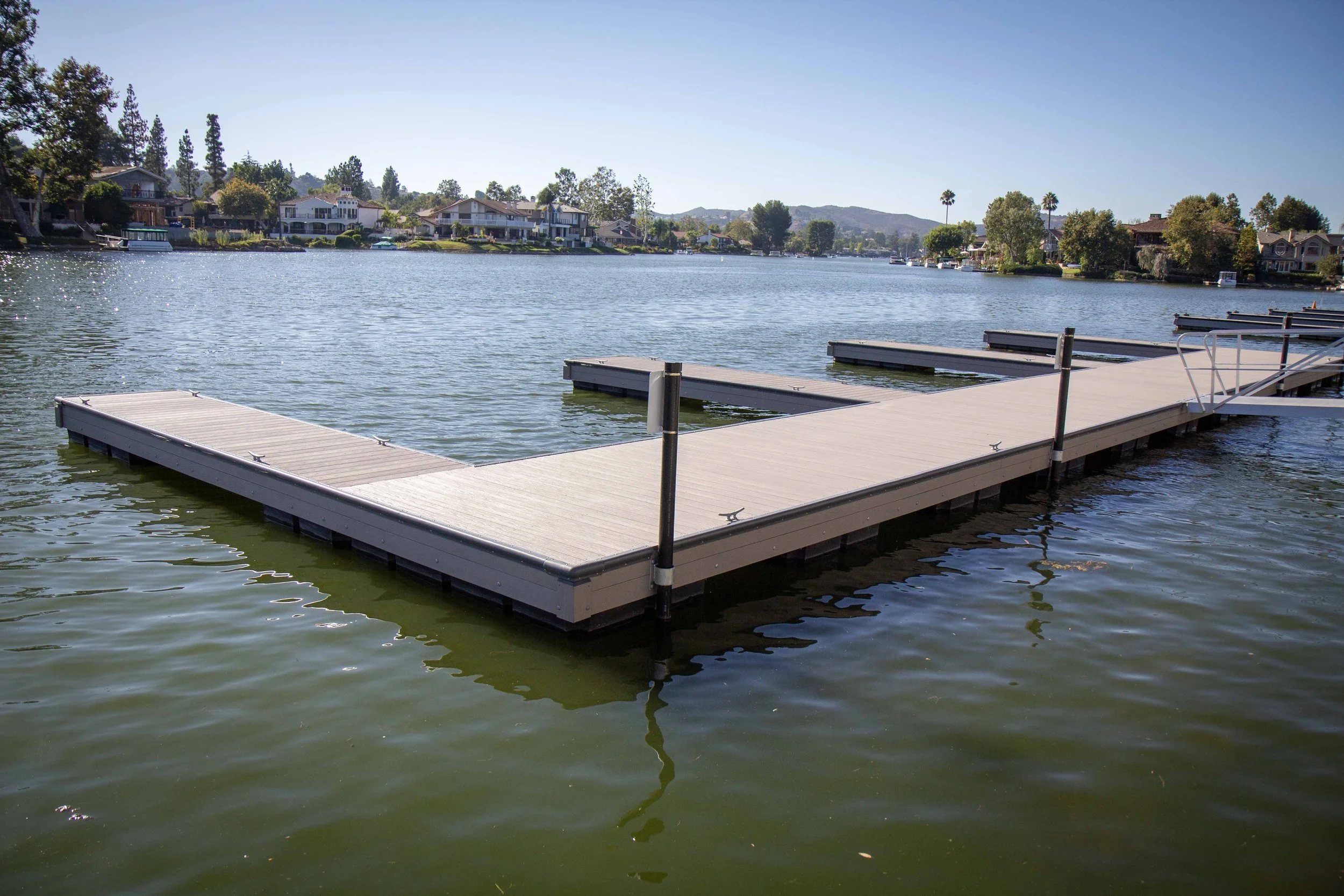 A floating dock extending on a calm lake with houses and trees in the background under a clear blue sky.