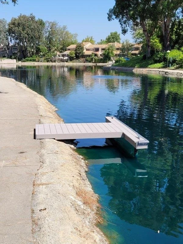 A small dock extends into a calm river with houses and trees in the background on a sunny day.