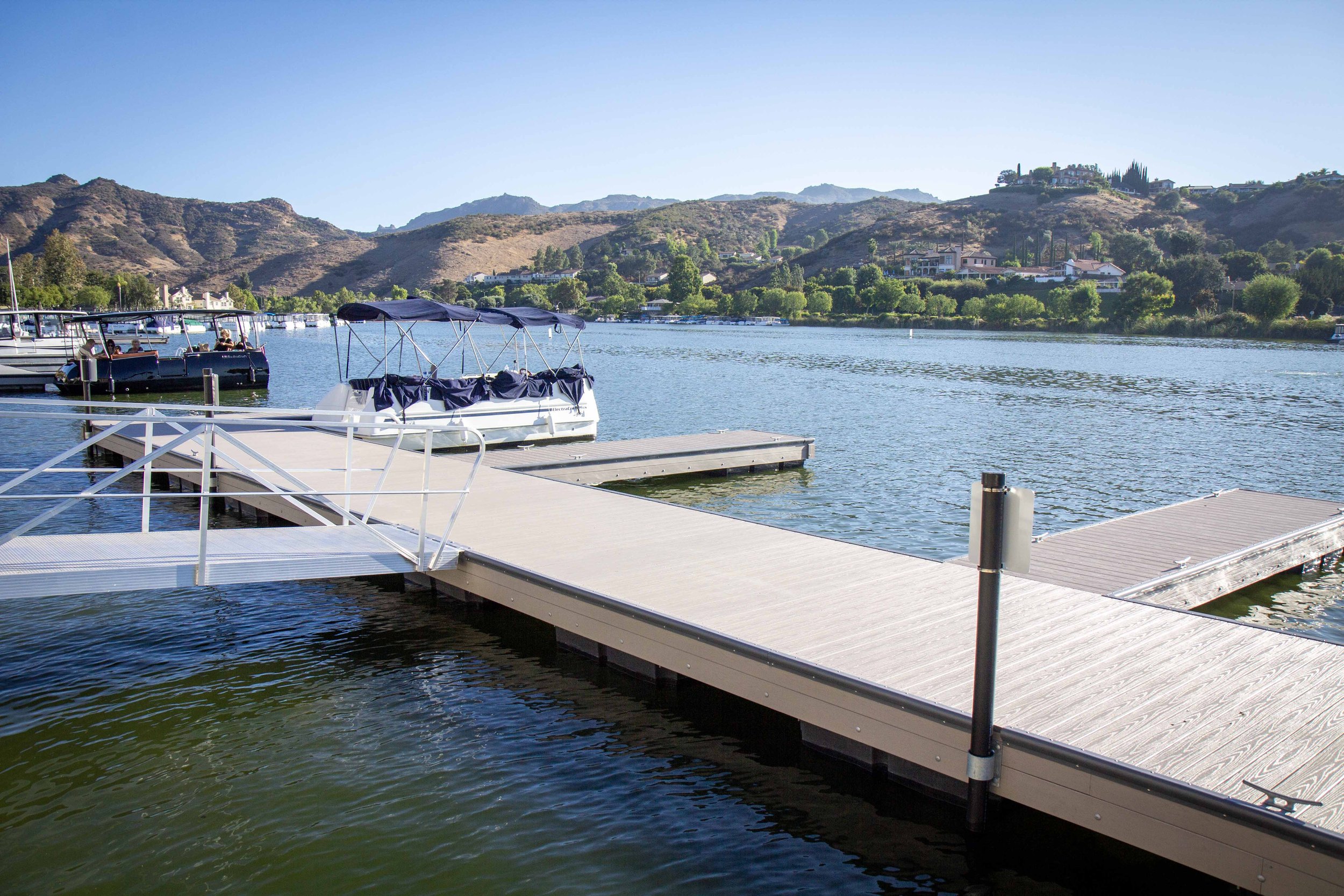 A marina with a paddle boat and multiple boats docked on the water, with hills and houses in the background on a clear day.