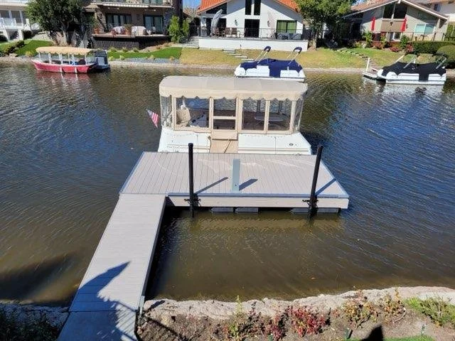 A motorboat docked at a waterway with houses and other boats in the background, including floating platforms and the shoreline with landscaped plants.