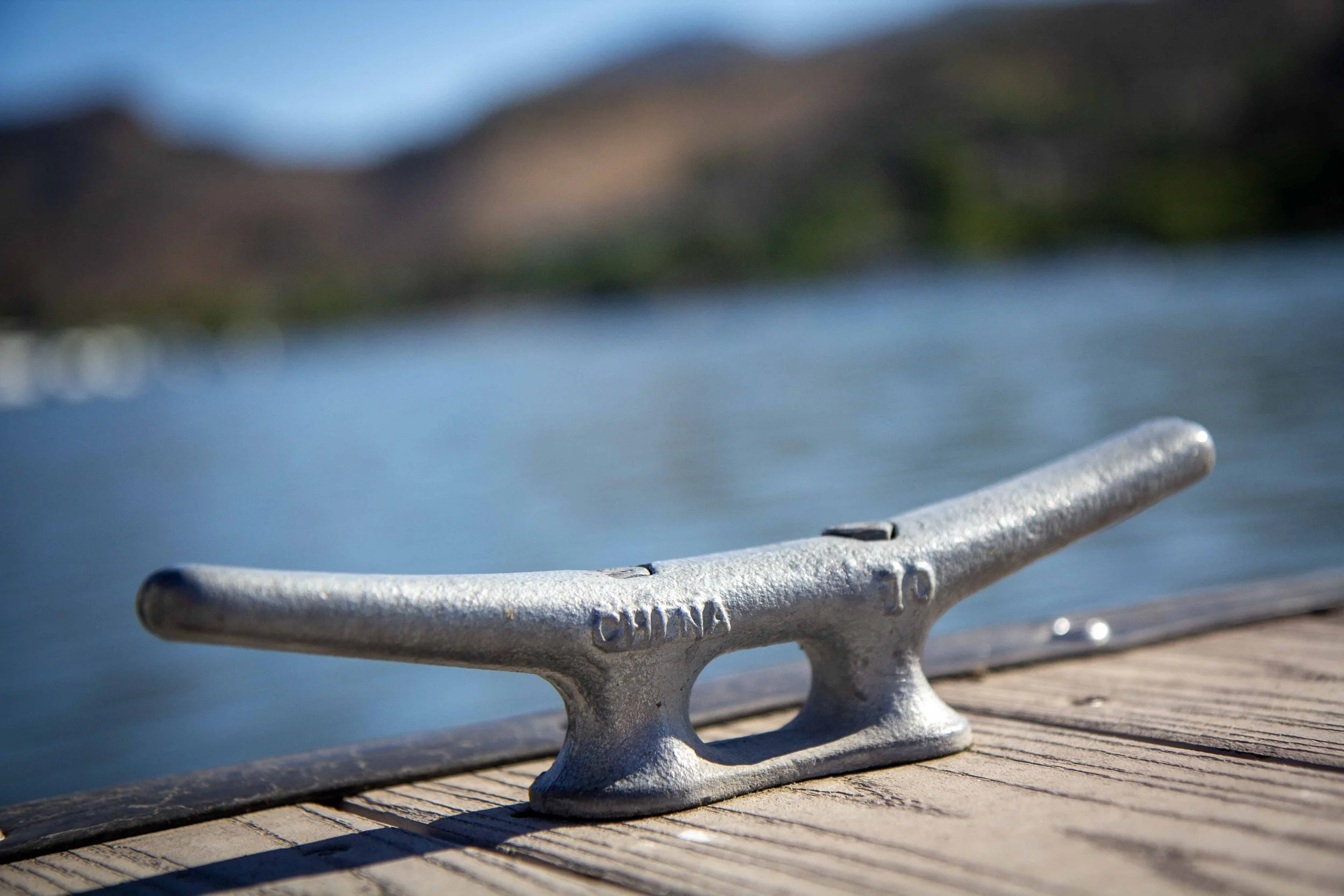 Close-up of a metal boat cleat attached to a wooden dock, with water and distant land in the background.