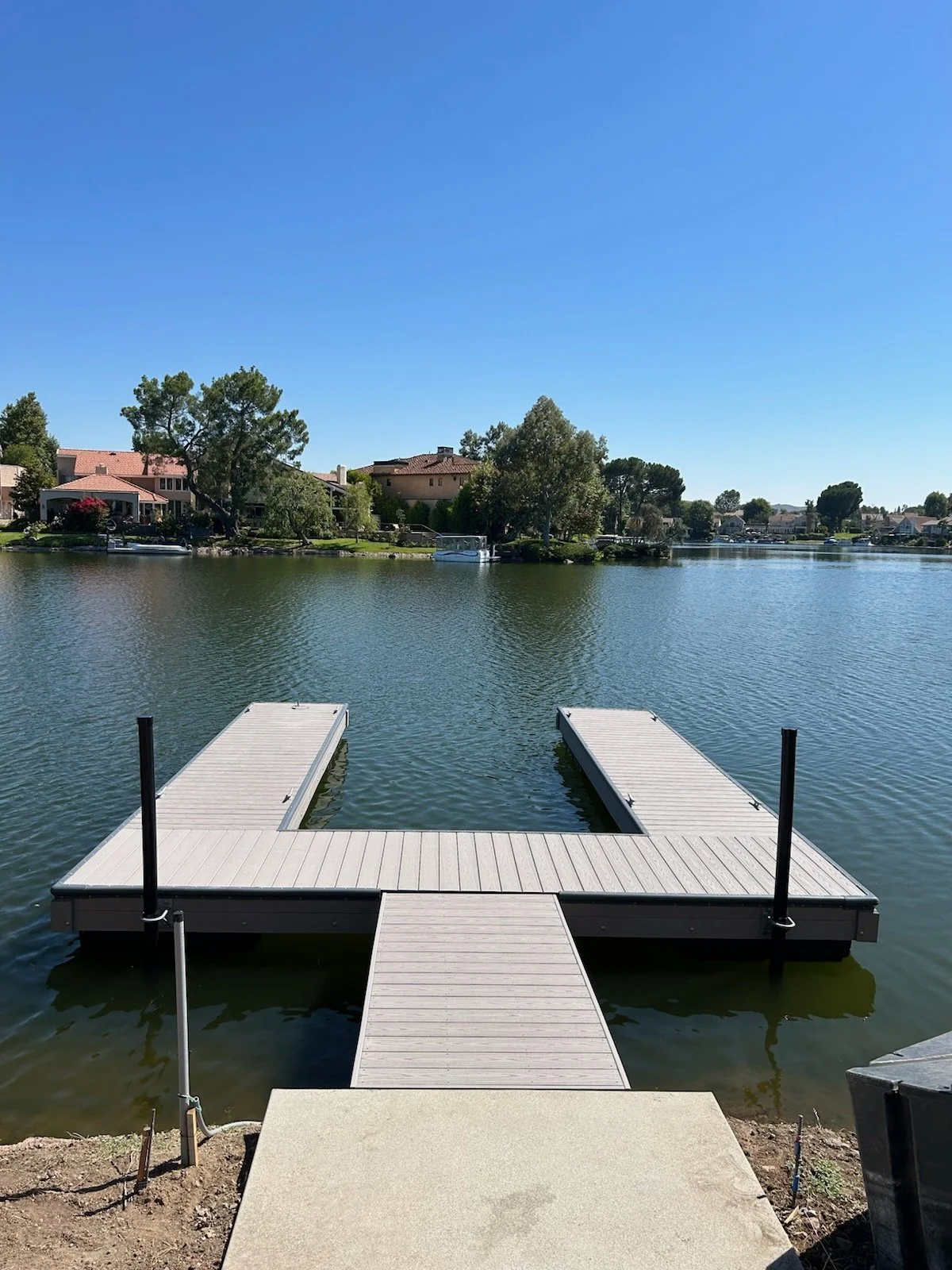 A calm lake with two empty boat docks extending into it, surrounded by residential houses and green trees under a clear blue sky.