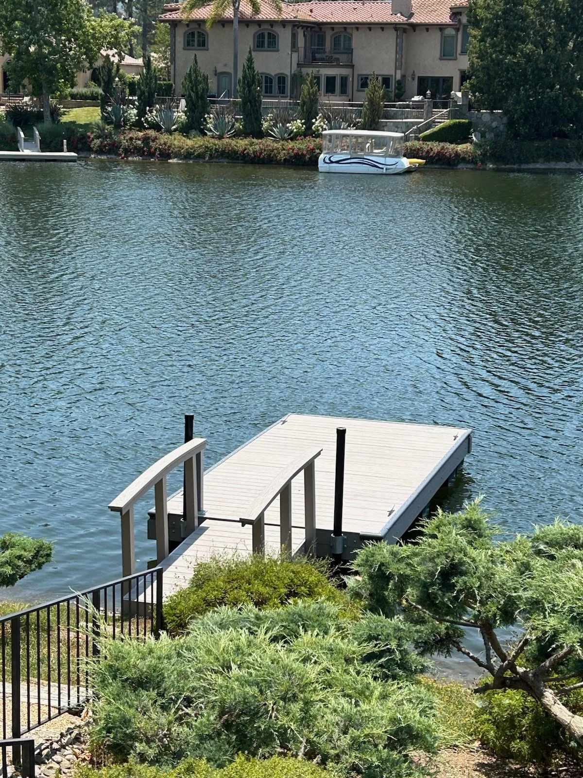 A small dock extends into a calm lake, with a white and blue paddle boat on the water nearby. A large house with multiple windows and balconies is visible in the background, surrounded by trees and landscaped gardens.