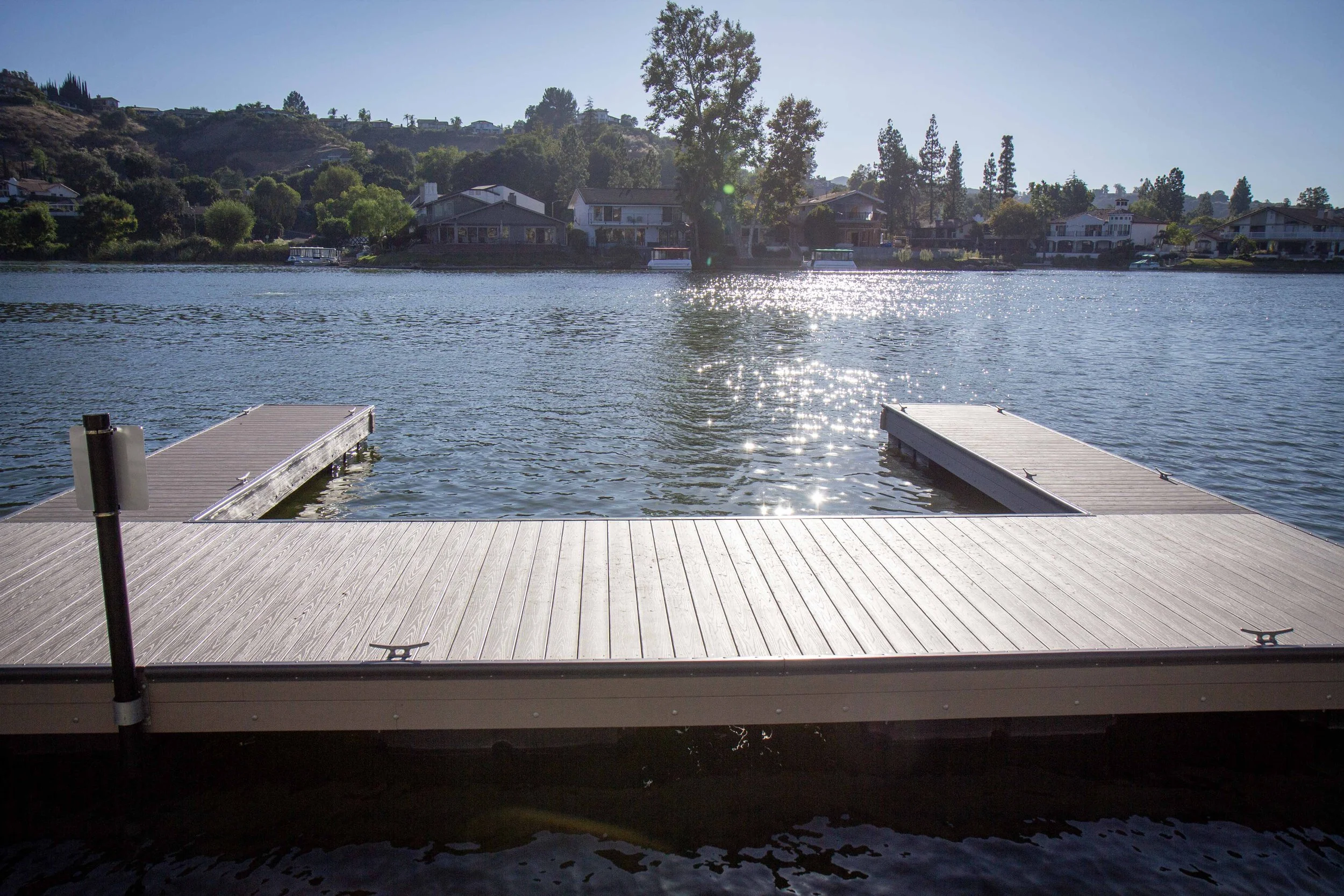 View of a wooden dock extending into a lake with houses and trees on the opposite shore. The sun is shining, creating reflections on the water.