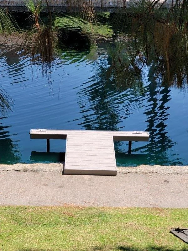 A small white dock extends into a calm, blue body of water, with overhanging trees reflected on the surface. A grassy area and concrete pathway are in the foreground.