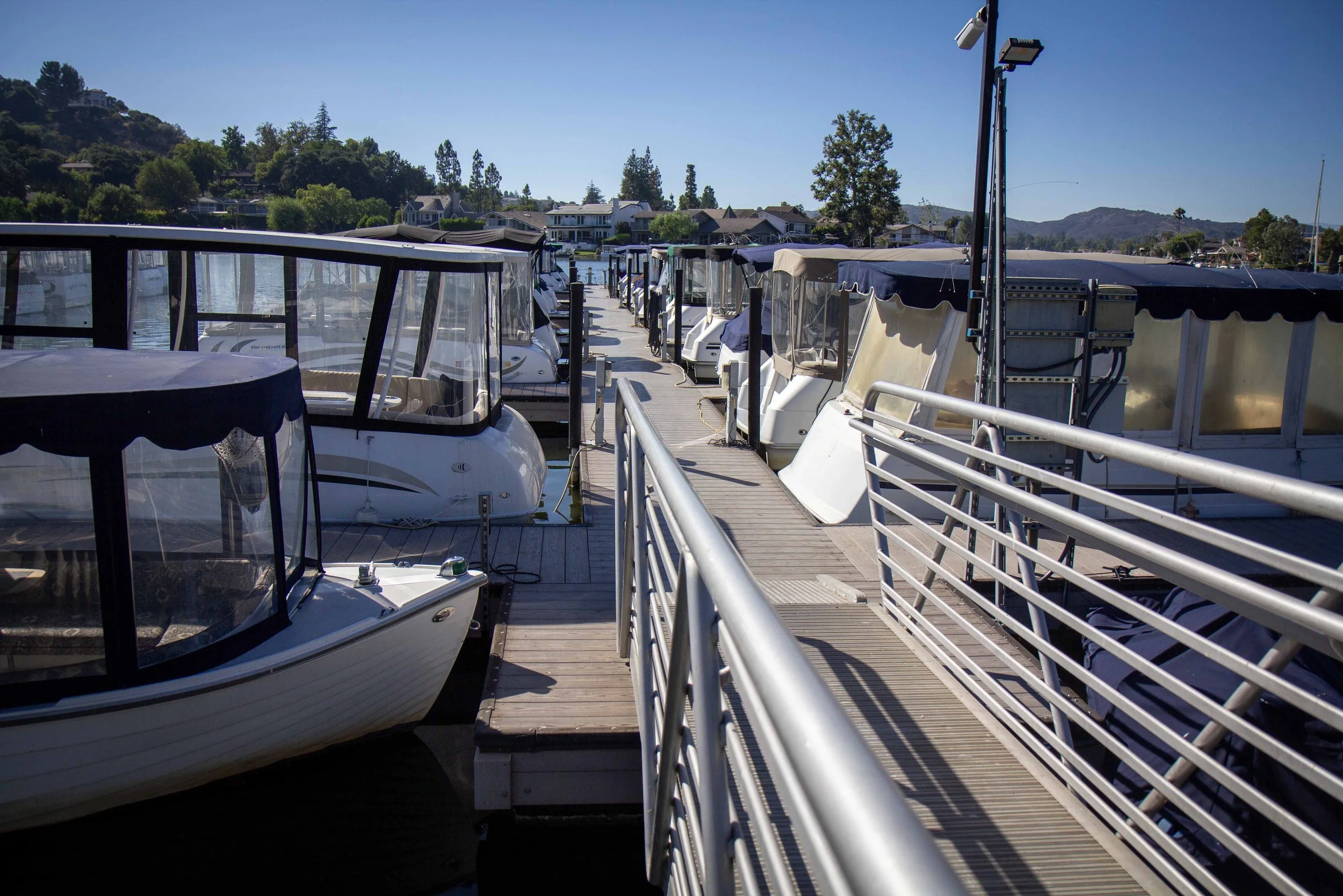 Boats docked at a marina with a floating wooden walkway, overlooking houses and hills under a clear blue sky.