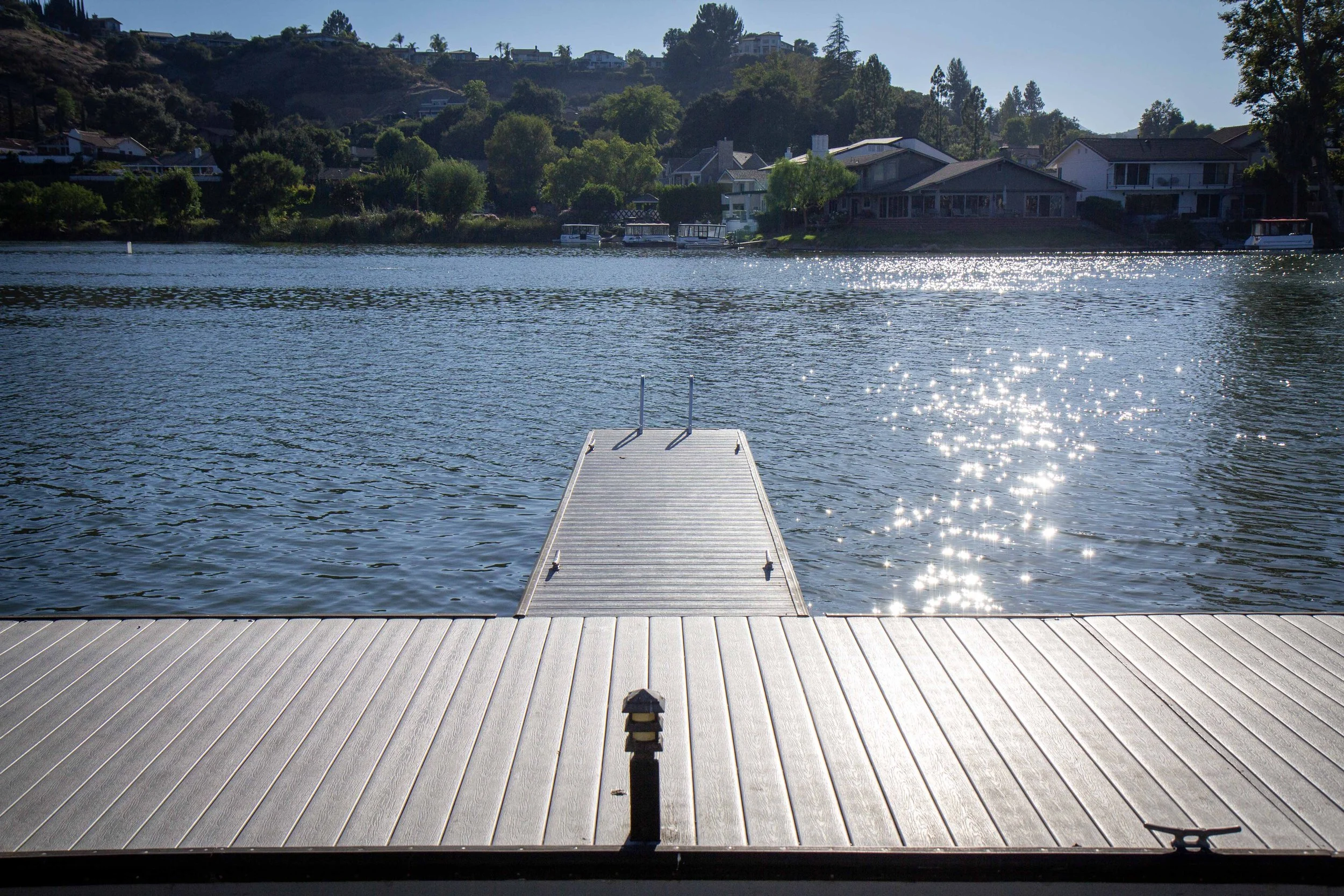 A wooden dock extends into a calm lake with sunlight reflecting off the water. In the background, houses are situated along a hillside with trees and a clear blue sky.