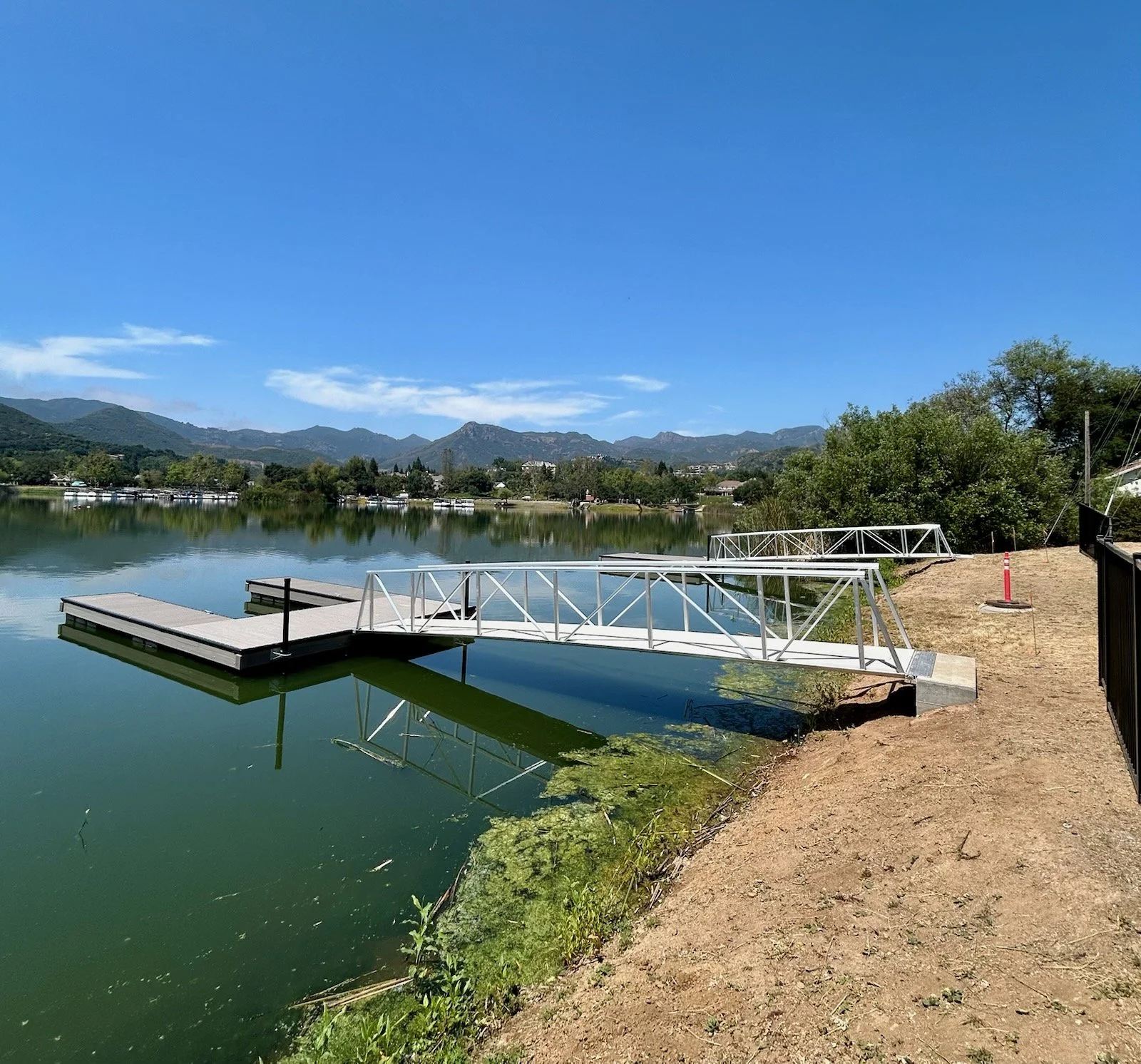 A dock extending into a calm lake with mountains and blue sky in the background, and some greenery around the shoreline.