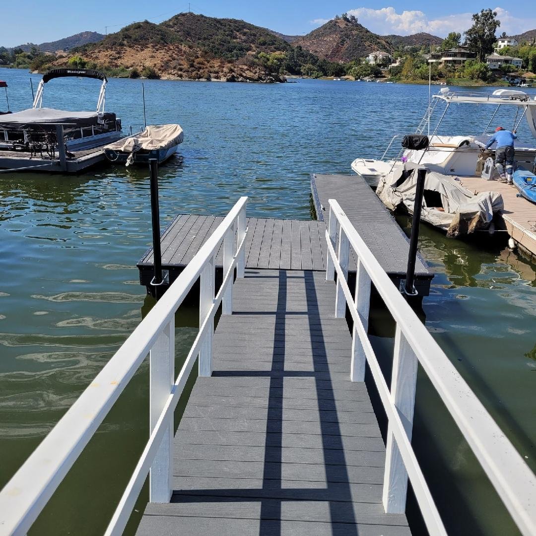 A small pier extending into a body of water with boats moored on either side, mountains and houses in the background, and partly cloudy sky.