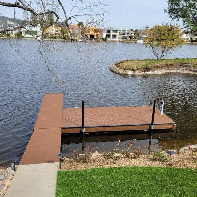 A lakeside backyard view with a brown wooden dock extending into the water, a small island with trees in the background, and residential houses across the lake, under a partly cloudy sky.