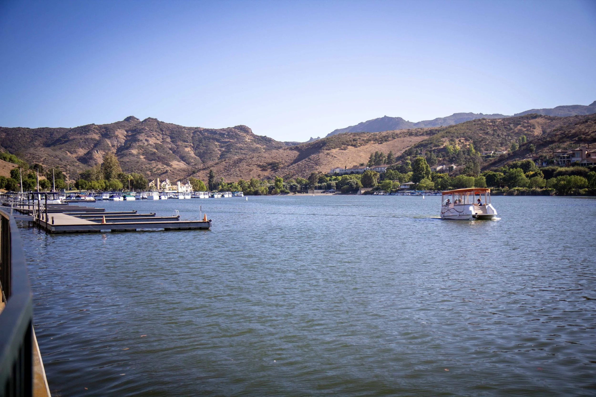 A peaceful body of water with a small boat cruising, surrounded by docked boats and rolling hills in the background under a clear blue sky.