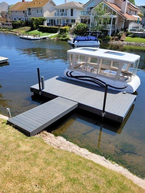 A white boat docked at a seaside patio with a gray ramp leading to a grassy yard, in front of colorful houses on a calm waterway.
