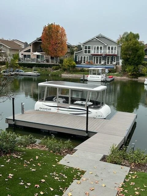 A dock with a boat moored on a calm river, residential houses in the background, and fall foliage on trees along the shoreline.