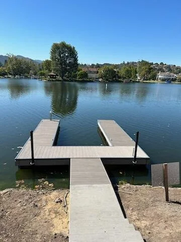 A small wooden dock extending into a calm lake with a clear blue sky above and trees in the background.
