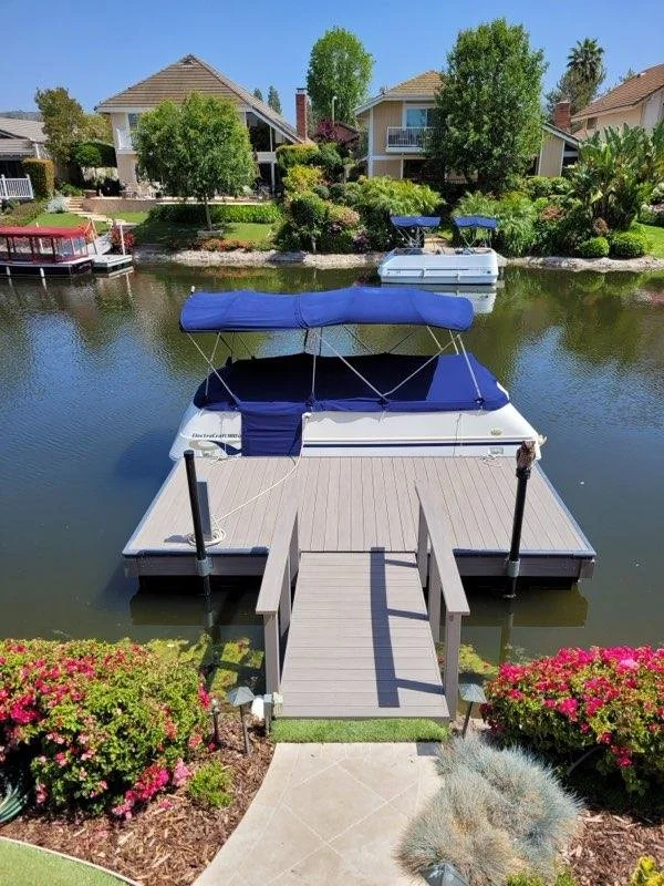 A boat dockside view from the backyard of a house, with a dock leading to a boat covered with a blue canopy docked along a calm waterway, with houses and trees in the background.