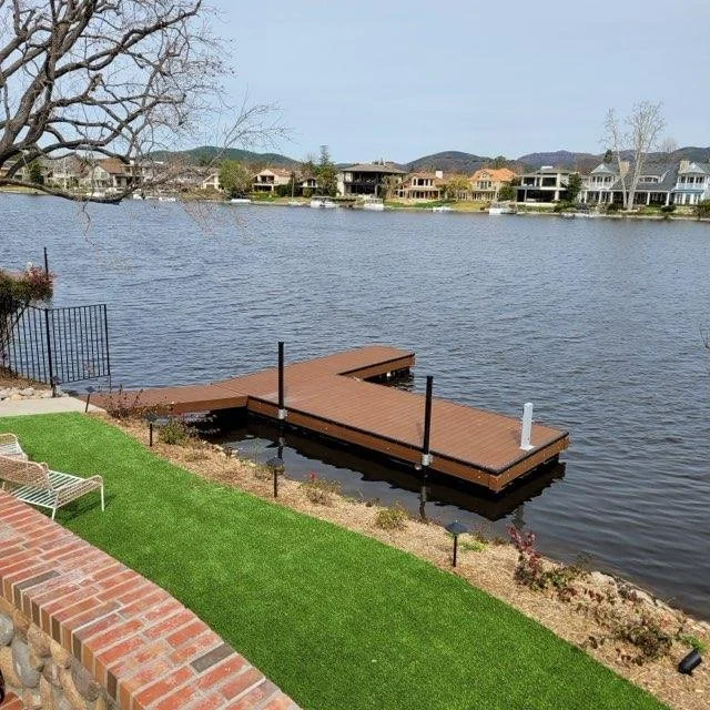 A lakeside backyard with a floating wooden dock, a green lawn with a brick border, and neighboring houses across the water.