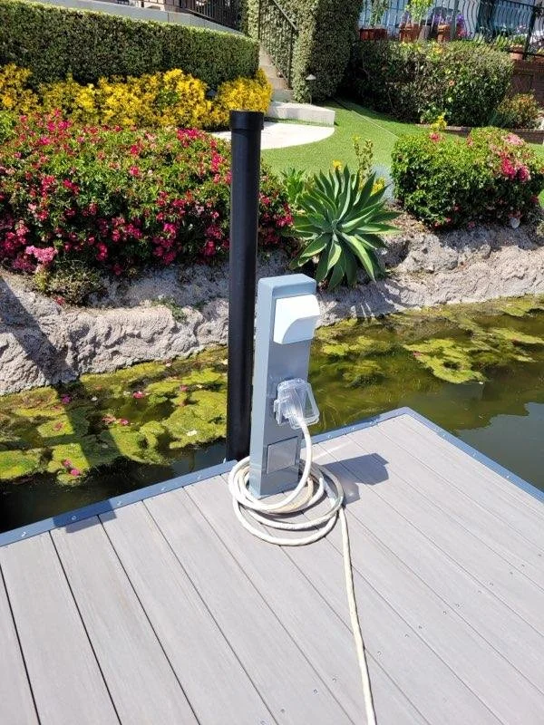 Electric vehicle charging station on a dock by a pond, with colorful flowers and garden in the background.