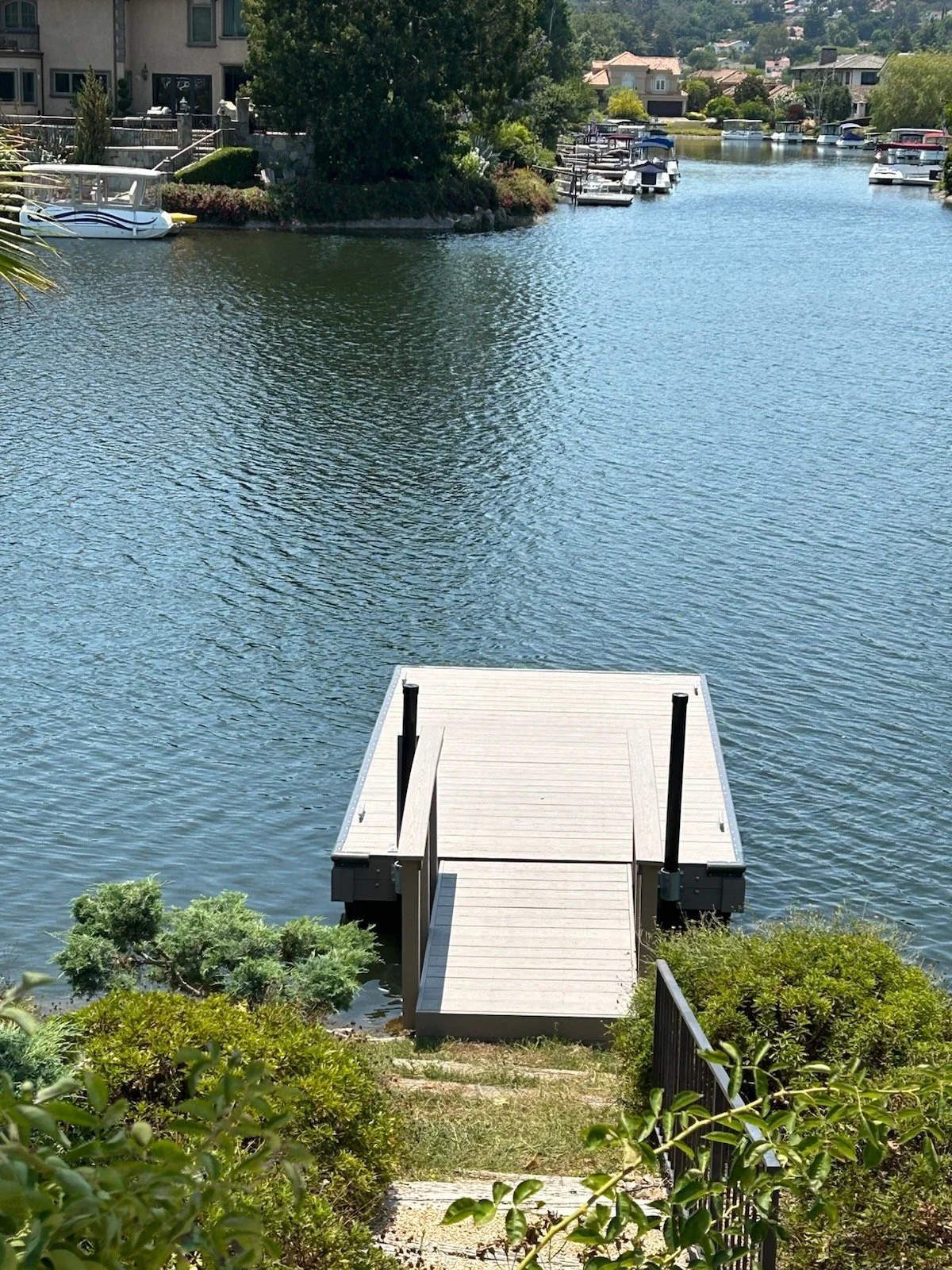 View of a small dock extending into a calm water canal with boats anchored on either side, surrounded by lush greenery and residential houses in the background.