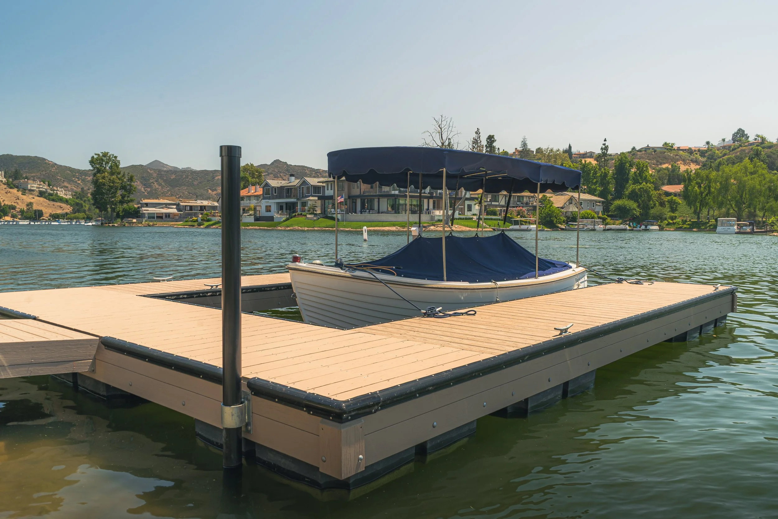 A boat docked at a wooden pier on a calm water body, with residential houses and green trees on a hillside in the background.