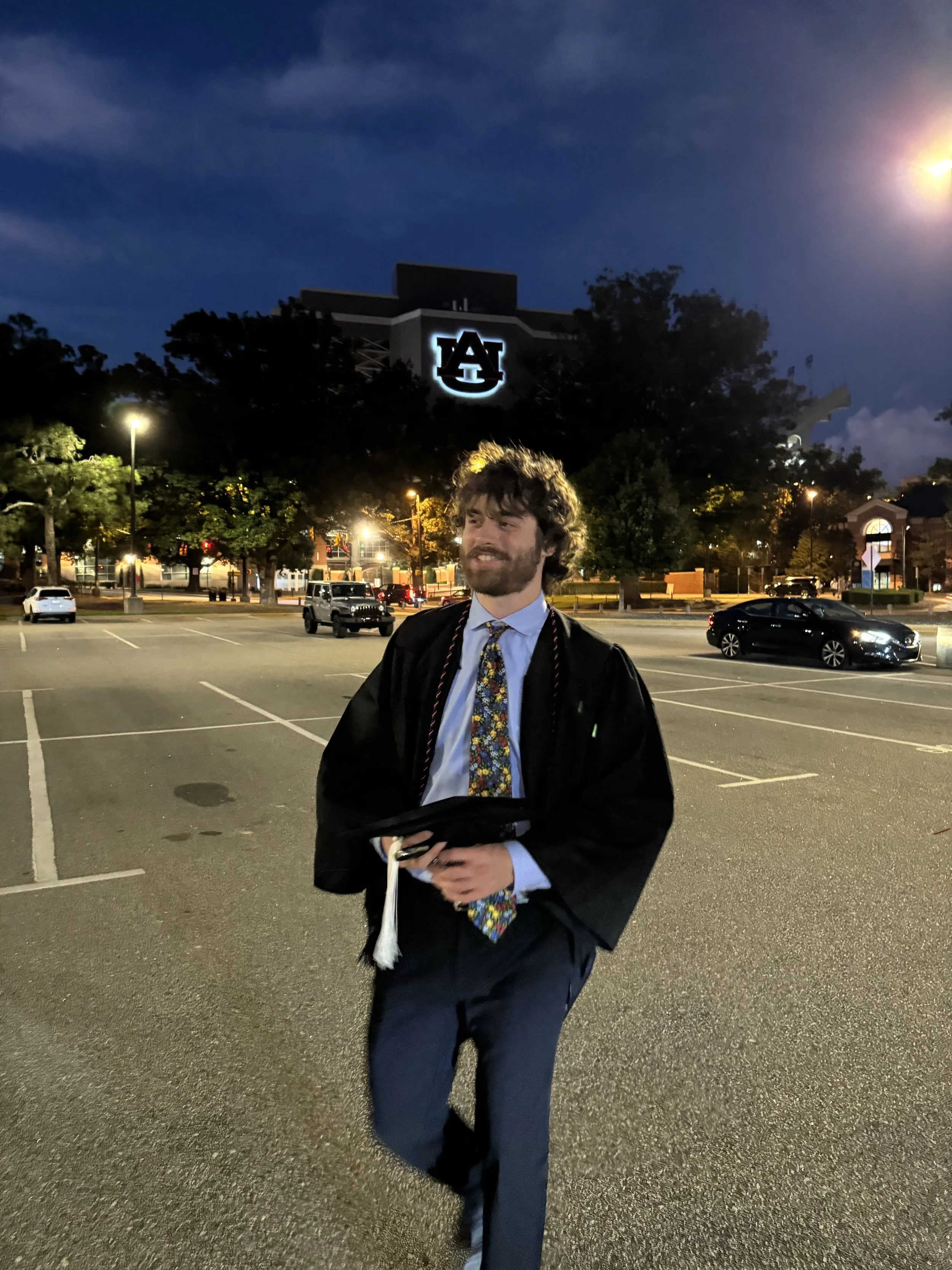 A man in graduation attire standing in an empty parking lot at night with illuminated university logo on a building in the background.