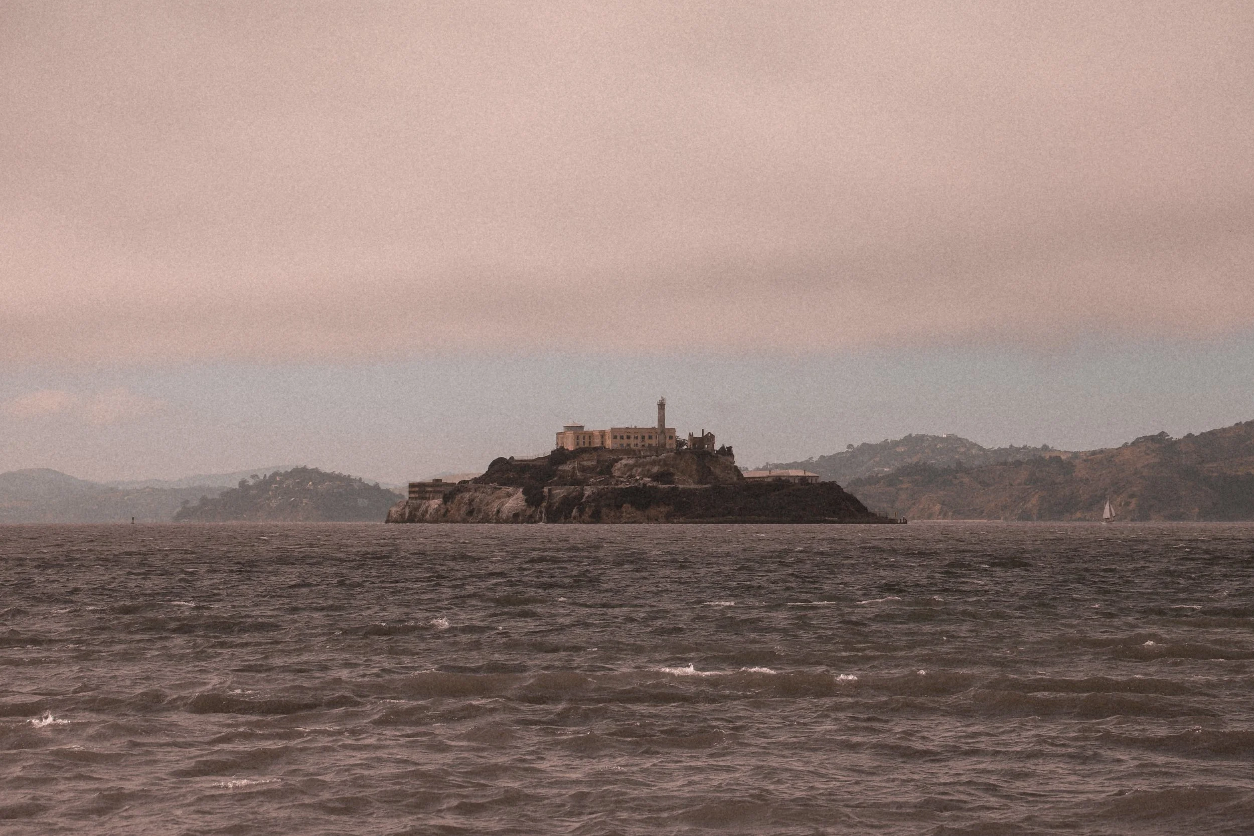 View of Alcatraz Island with a lighthouse on a rocky terrain, surrounded by water, and hills in the background under a pale pinkish sky.