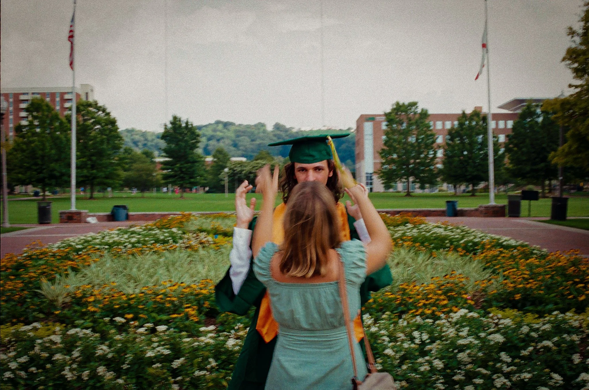 A young man in a green graduation cap and gown smiling as a young woman adjusts his cap outdoors on a college campus with flowers and trees.