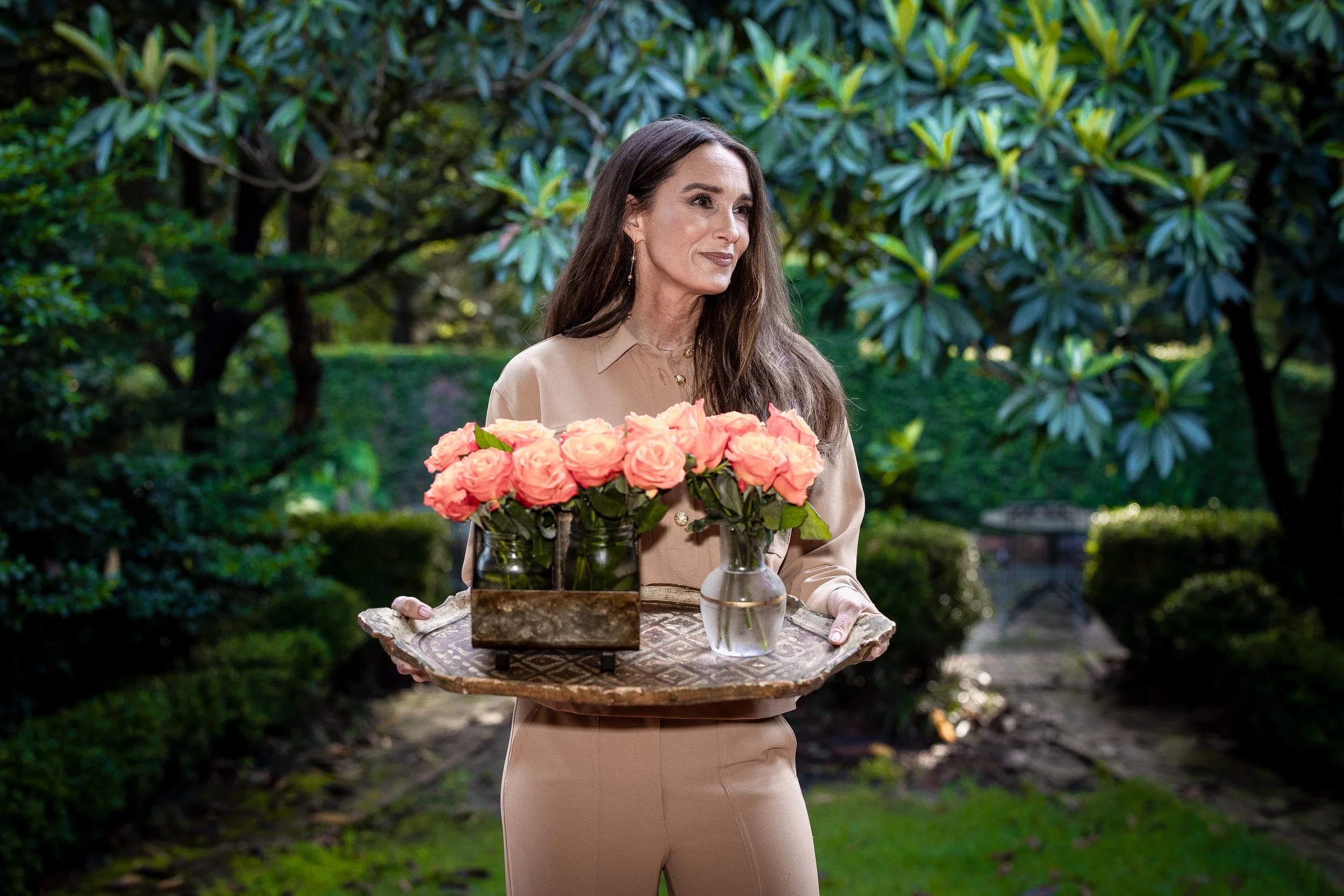A woman with long dark hair, dressed in beige, standing outdoors in a lush garden, holding a tray with three glasses of peach-colored roses in vases.