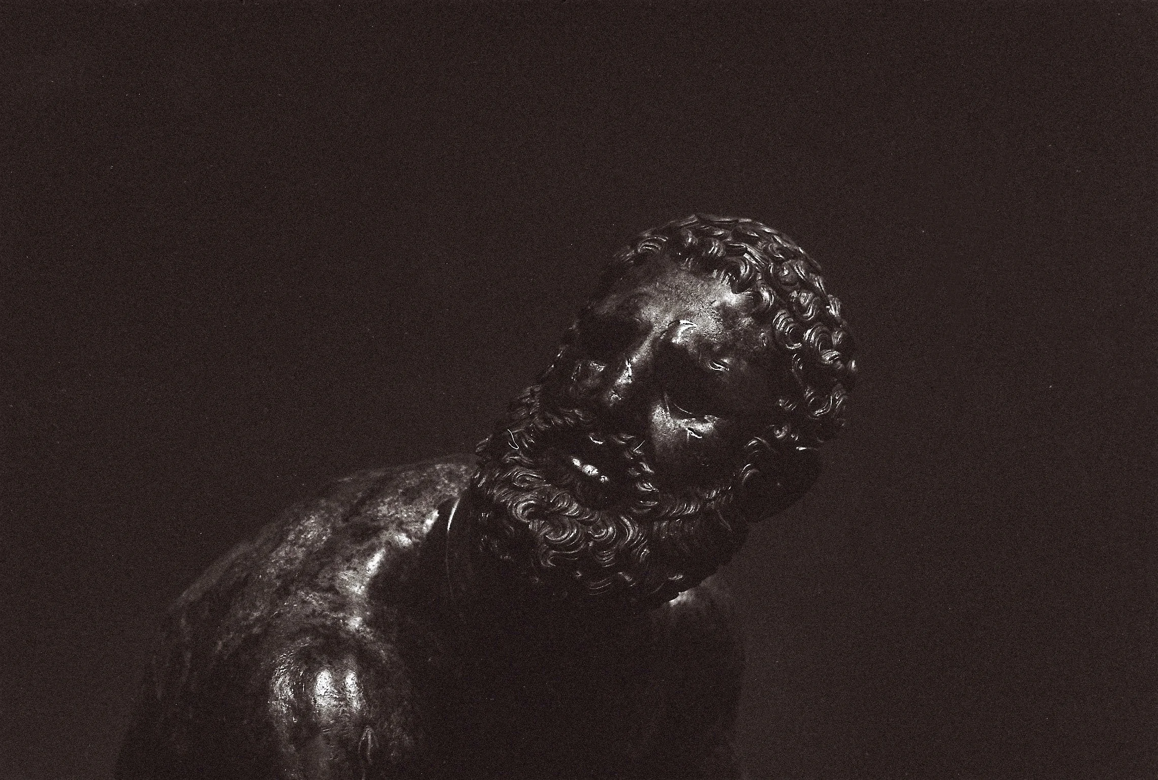 Close-up of a bronze bust sculpture of a bearded man with curly hair, against a dark background.