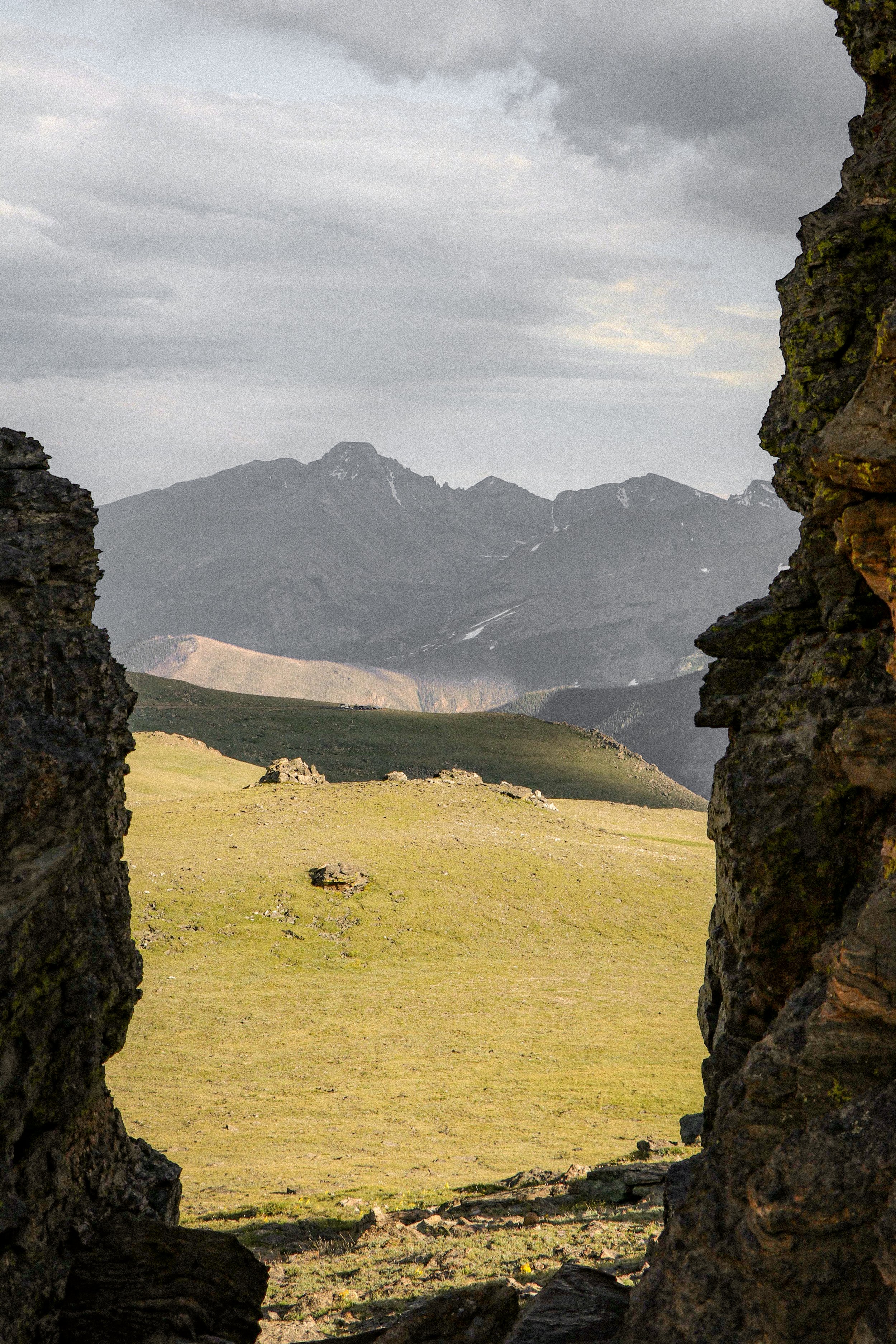 View of mountains through a rocky opening, with grassy terrain in the foreground and cloudy sky above.