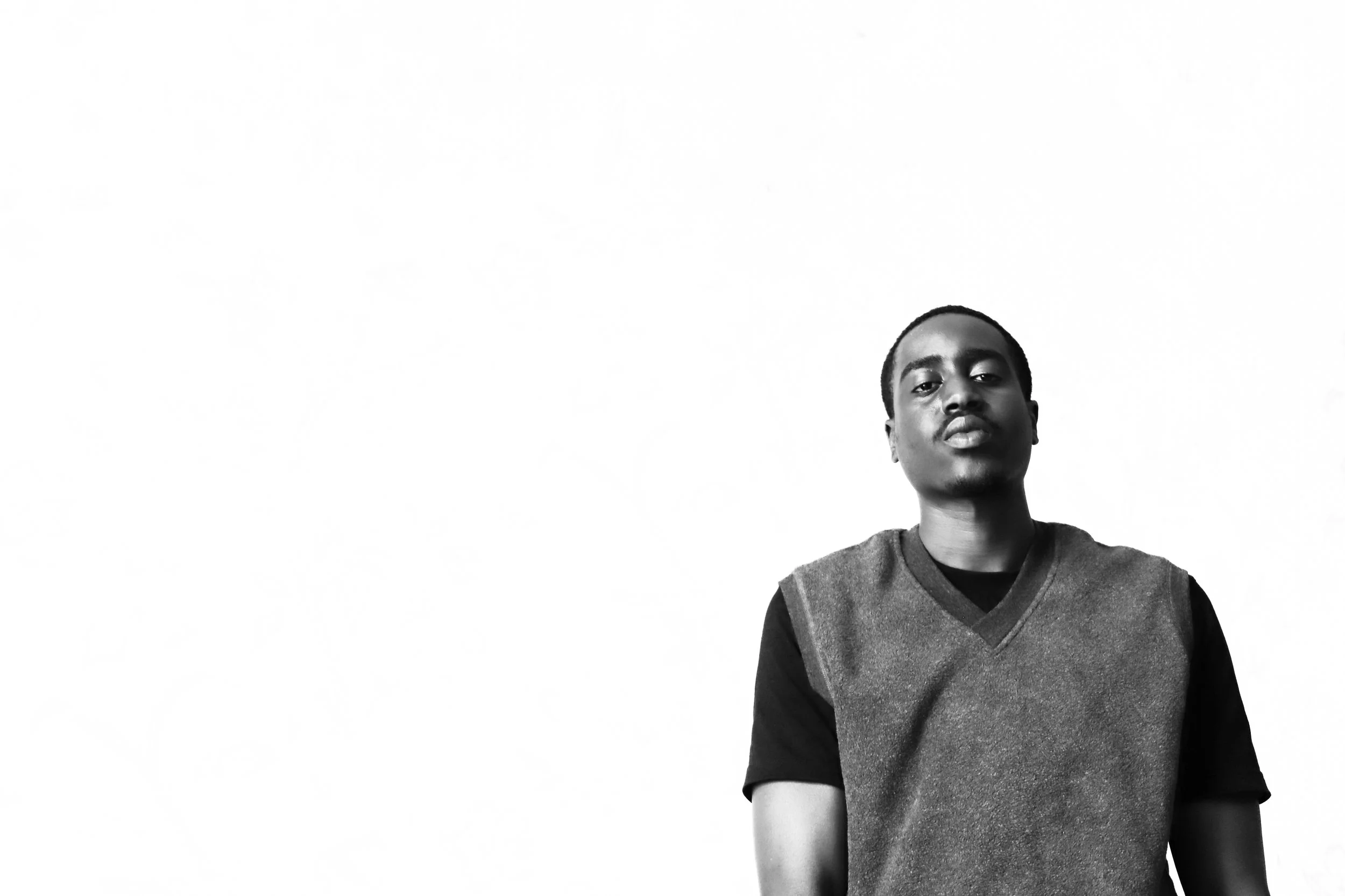 A  man wearing a dark t-shirt and a sleeveless vest, standing against a plain white background, facing the camera with a serious expression.