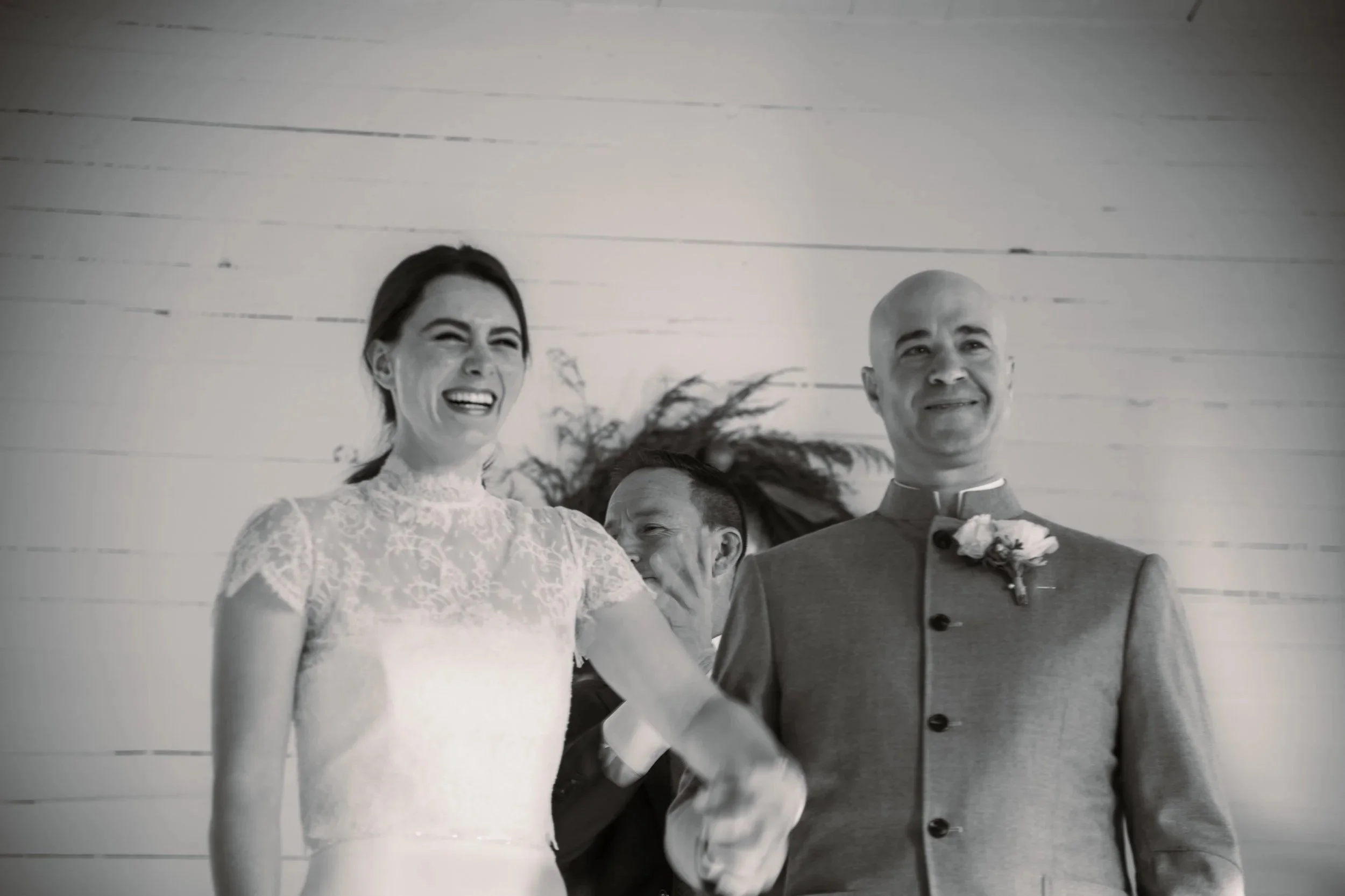 A bride and groom holding hands during their wedding ceremony, laughing and smiling, with a man in the background smiling, all in front of a light-colored wooden wall.